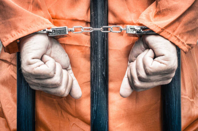 shutterstock_217094626.jpg featured image Handcuffed person in orange jumpsuit behind prison bars.