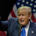 Man pointing while speaking at podium with American flags.