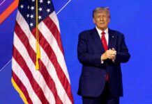 Donald Trump in suit near the American flag on stage.