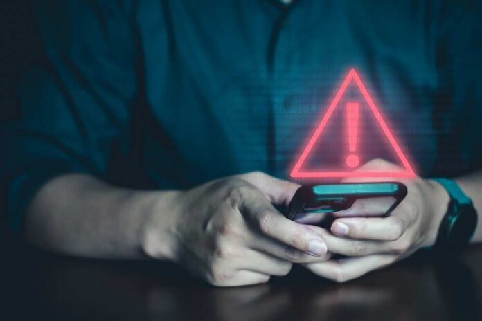 shutterstock_2342619545.jpg featured image Person holding phone with warning symbol displayed.