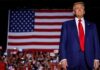 Donald Trump in suit stands before large American flag