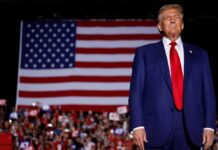 Donald Trump in suit stands before large American flag