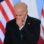 Joe Biden coughing in a suit standing in front of flags.