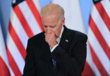 Joe Biden coughing in a suit standing in front of flags.