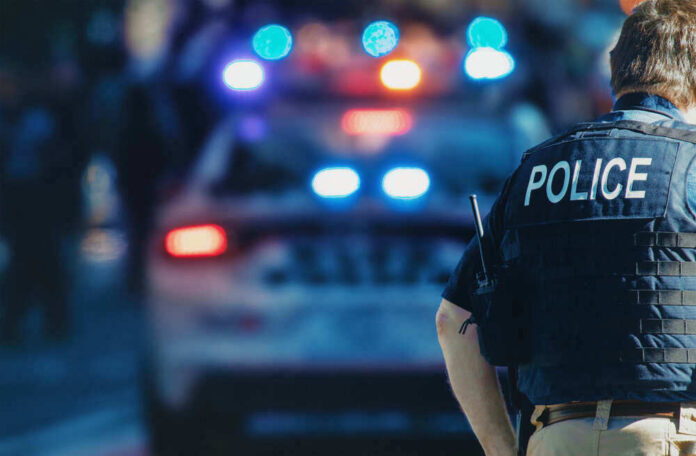 shutterstock_1543591088-1.jpg Police officer stands near patrol car with lights flashing
