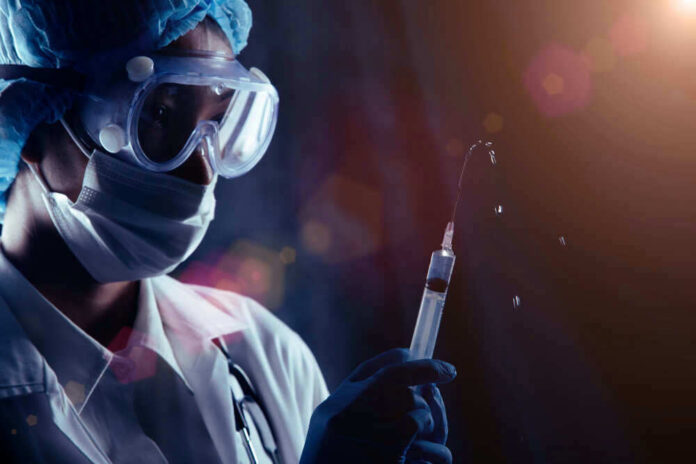 Person in PPE holding syringe with liquid droplets.