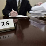 IRS sign on desk, person handling paperwork behind.