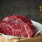 A raw cut of beef on a wooden board with herbs and a pepper grinder