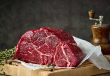 A raw cut of beef on a wooden board with herbs and a pepper grinder