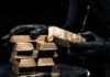 CIA Gold Scandal Exposed A person in black gloves handling gold bars against a dark background