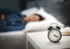 House Gambles With Sleep — Health Warning Ignored? An alarm clock on a bedside table with a person sleeping in the background
