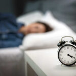 An alarm clock on a bedside table with a person sleeping in the background