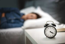 An alarm clock on a bedside table with a person sleeping in the background