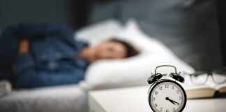 An alarm clock on a bedside table with a person sleeping in the background