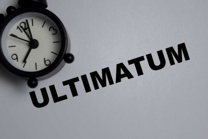 Concept,Of,Ultimatum,Write,On,Book,Isolated,On,Wooden,Table. A black alarm clock next to the word 'ULTIMATUM' on a gray background