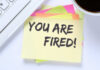 A sticky note with the message 'YOU ARE FIRED!' placed on a desk next to a keyboard and a cup of coffee