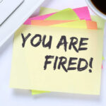 A sticky note with the message 'YOU ARE FIRED!' placed on a desk next to a keyboard and a cup of coffee