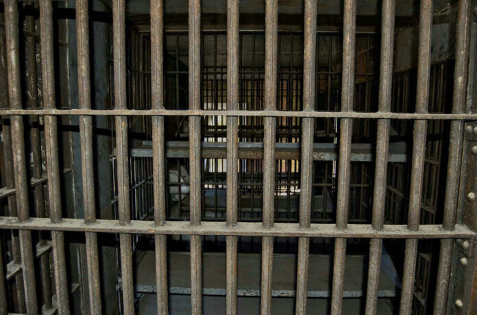 shutterstock_747318307 Empty, rusty barred jail cell with metal bunks.