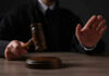 Judge holding gavel, hand raised in courtroom.