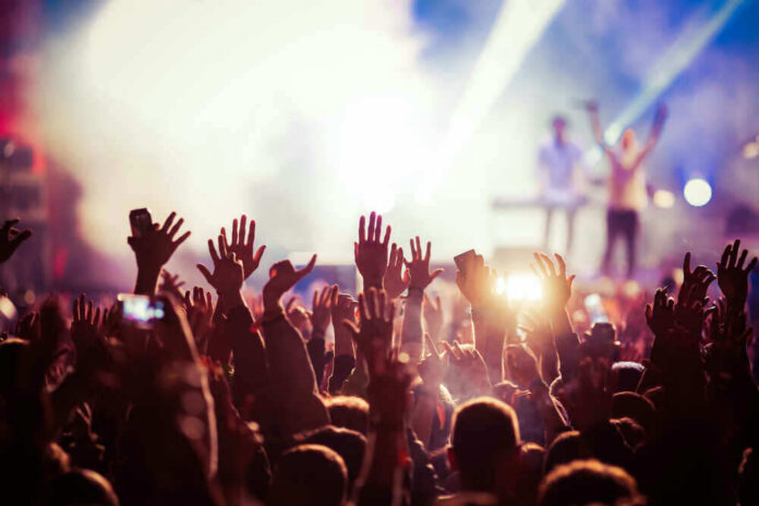 shutterstock_611045375 Concert crowd with hands raised, bright stage lights.
