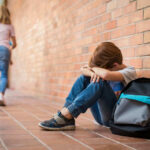 Sad child sitting against brick wall, kids running away.