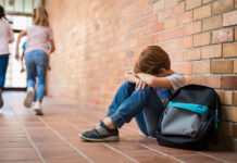 Sad child sitting against brick wall, kids running away.