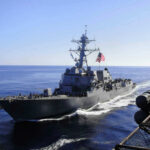 U.S. Strikes Iran Amid Fragile Ceasefire Warship with American flag sailing in the ocean.