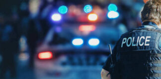 Police officer stands near patrol car with lights flashing.