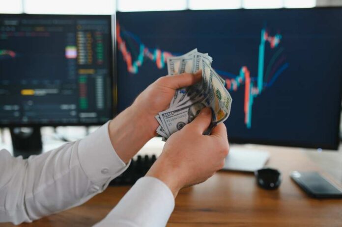 Person holding cash in front of stock market charts on computer screens