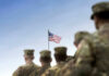 Soldiers in uniform facing American flag outdoors.
