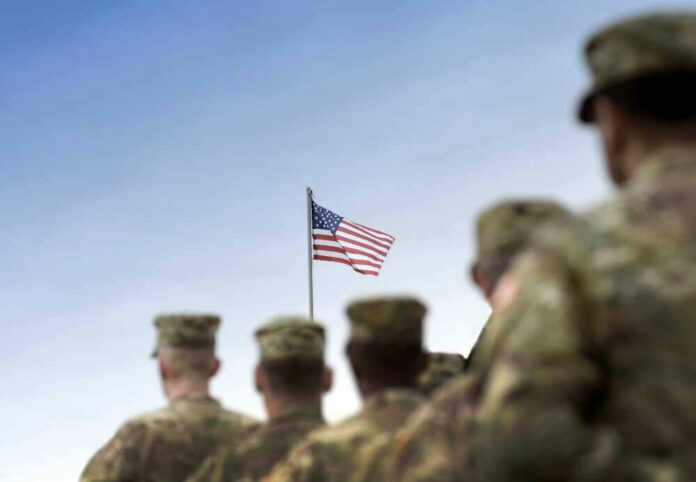 shutterstock_2380883205 Soldiers in uniform facing American flag outdoors.