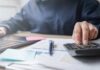 Paychecks Hit As THESE Costs Surge Again Person using a calculator while working on a laptop and reviewing documents