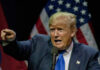 Man pointing while speaking at podium with American flags.