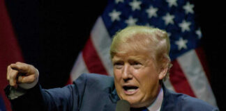 Man pointing while speaking at podium with American flags.